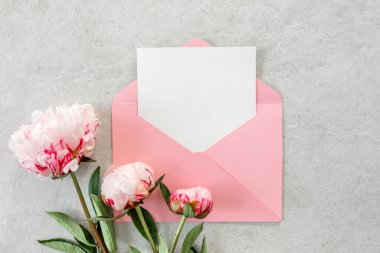 Sahte davetiye, boş kağıt tebrik kartı, pembe zarf ve gri taş masada şakayıklar. Çiçek arkaplanı. Düz yatış, üst görünüm.