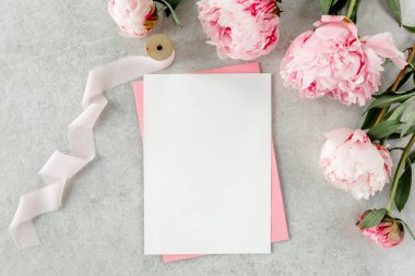 Sahte davetiye, boş kağıt tebrik kartı, pembe zarf ve gri taş masada şakayıklar. Çiçek arkaplanı. Düz yatış, üst görünüm.