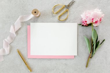 Sahte davetiye, boş kağıt tebrik kartı, pembe zarf ve gri taş masada şakayıklar. Çiçek arkaplanı. Düz yatış, üst görünüm.