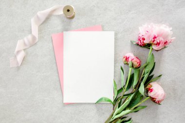 Sahte davetiye, boş kağıt tebrik kartı, pembe zarf ve gri taş masada şakayıklar. Çiçek arkaplanı. Düz yatış, üst görünüm.