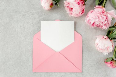 Sahte davetiye, boş kağıt tebrik kartı, pembe zarf ve gri taş masada şakayıklar. Çiçek arkaplanı. Düz yatış, üst görünüm.