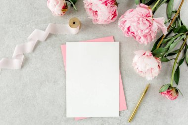 Sahte davetiye, boş kağıt tebrik kartı, pembe zarf ve gri taş masada şakayıklar. Çiçek arkaplanı. Düz yatış, üst görünüm.
