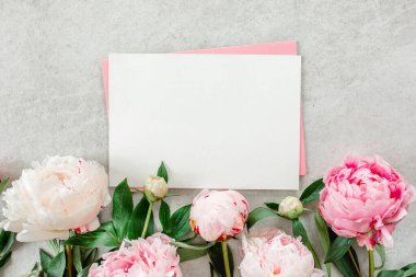 Sahte davetiye, boş kağıt tebrik kartı, pembe zarf ve gri taş masada şakayıklar. Çiçek arkaplanı. Düz yatış, üst görünüm.