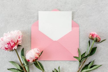 Sahte davetiye, boş kağıt tebrik kartı, pembe zarf ve gri taş masada şakayıklar. Çiçek arkaplanı. Düz yatış, üst görünüm.
