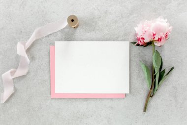 Sahte davetiye, boş kağıt tebrik kartı, pembe zarf ve gri taş masada şakayıklar. Çiçek arkaplanı. Düz yatış, üst görünüm.
