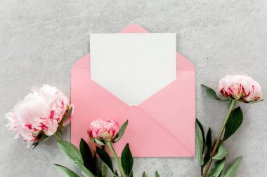 Sahte davetiye, boş kağıt tebrik kartı, pembe zarf ve gri taş masada şakayıklar. Çiçek arkaplanı. Düz yatış, üst görünüm.