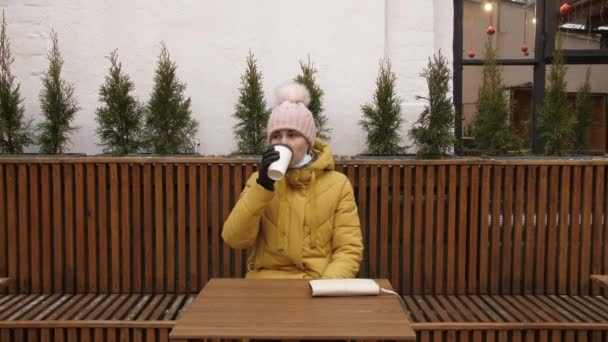 Портрет молодой девушки в пальто, сидящей на улице в пустом кафе. Расслабляющая женщина с кофе, чтобы пойти. — стоковое видео