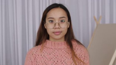 Kendine güveni tam, gözlüklü genç Asyalı kadın ofiste kameraya bakıyor. Güzel bir kadın, kapalı alanda yakın plan portre pozu veriyor. Yavaş çekim.