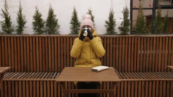 Junge Frau trinkt Kaffee in einem Straßencafé, frostiger Morgen vor dem Arbeitstag, genießt Morgenkaffee, Loft-Café mit Kaffee zum Mitnehmen, Zeitlupe — Stockvideo