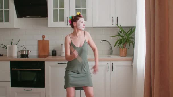 Una joven se divierte y baila, canta en un cucharón, ve videos musicales, escucha música en una cocina moderna, cocina comida en casa, se cuida. — Vídeo de stock