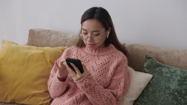 美しいアジアの若い女性がスマートフォンでリビングルームのソファに座って、インターネットスーパーマーケット、オンライン、配達、食品、注文でオンラインで購入します。セルガジェット技術. — ストック動画