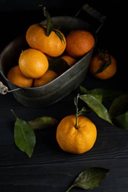 Konteynırda yaprakları olan birkaç mandalina siyah bir masanın üzerinde koyu bir zemin üzerinde yatıyor. Rustik biçim kopyalama uzayı.