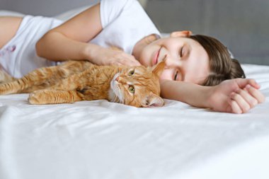 Kızıl kedili bir kız beyaz bir yatakta yatıyor. Genç bir kız beyaz bir çarşafta yatarken kızıl bir kediyi okşuyor.