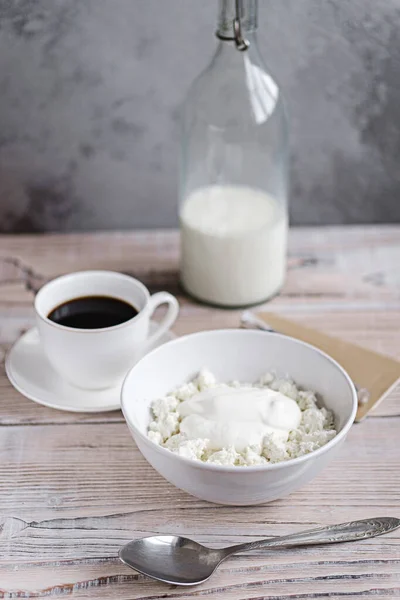 Queso de cabaña con crema agria en un tazón blanco con café y leche en una mesa de madera blanca. Desayuno saludable. Estilo rústico Imagen de stock