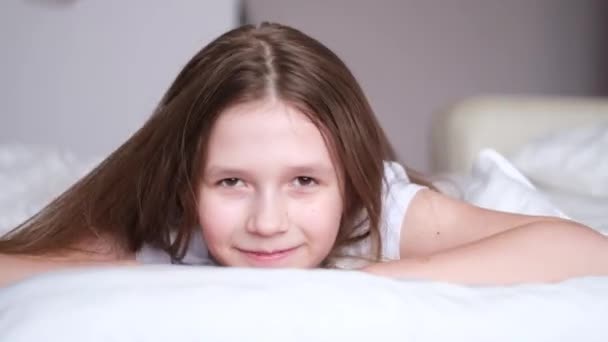 7-9岁的小女孩，穿着白色T恤，躺在白色的床上，微笑着环顾四周。4K视频。四.后续行动. — 图库视频影像