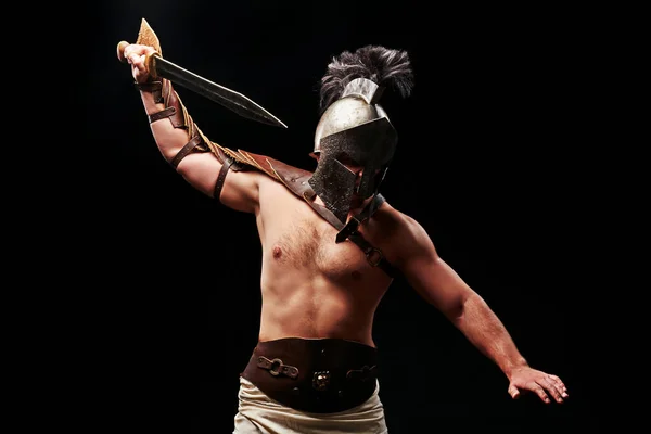 Gladiator with sword and armor on a black background. A warrior in gladiatorial armor and a helmet cuts with a sword in front of him. — Stock Photo, Image