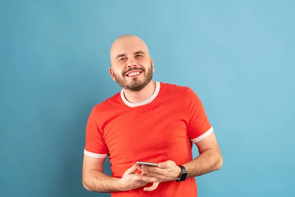 Ein glatzköpfiger Mann mittleren Alters mit Bart und rotem T-Shirt auf blauem Grund. Hält ein Telefon in der Hand und zeigt mit dem Finger der anderen Hand darauf. — Stockfoto