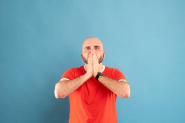 Mavi arka planda sakallı ve kırmızı tişörtlü orta yaşlı kel bir adam, korkuyla yüzünü elleriyle kapatıyor..