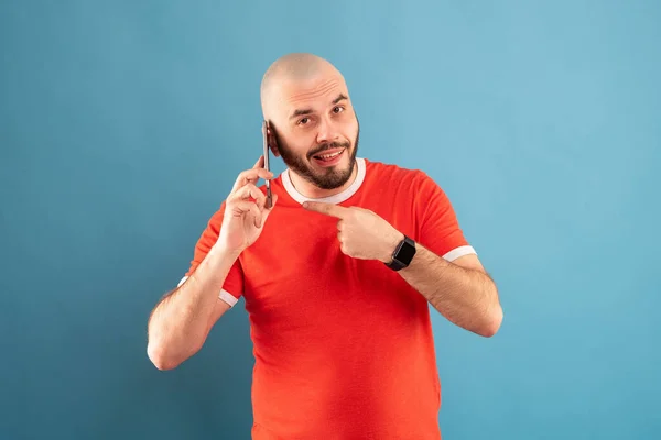 Ein glatzköpfiger Mann mittleren Alters mit Bart und rotem T-Shirt auf blauem Grund. Hält ein Telefon in der Hand und zeigt mit dem Finger der anderen Hand darauf — Stockfoto