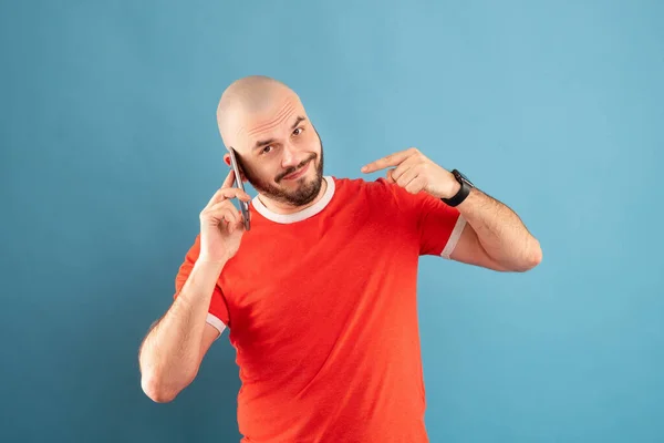 Ein glatzköpfiger Mann mittleren Alters mit Bart und rotem T-Shirt auf blauem Grund. Hält ein Telefon in der Hand und zeigt mit dem Finger der anderen Hand darauf — Stockfoto