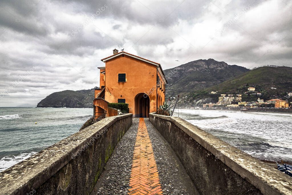 Symmetrical view of an italian brick bridge and pier with an orange house against rough sea and a cloudy sky near five towns, Levanto, Liguria, La Spezia, Italy