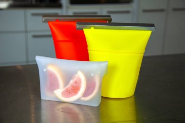 Üç tane sıfır atıklı silikon yiyecek torbası, atıştırmalıkları tutmak için ya da plastik zip-kilit tarzı çantaların yerine kurutulmuş ürünleri koymak için. Kırmızı ve sarı bir çanta greyfurtlu açık büfenin yanında duruyor..