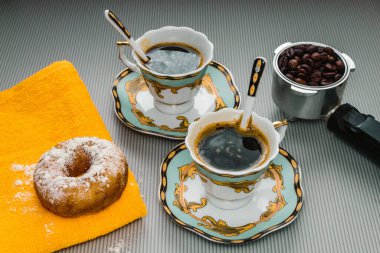 Sarı masa örtüsünün üzerinde kızarmış çörek, klasik kahve seti ve kahve kornası var. Açık çizgili arkaplan.