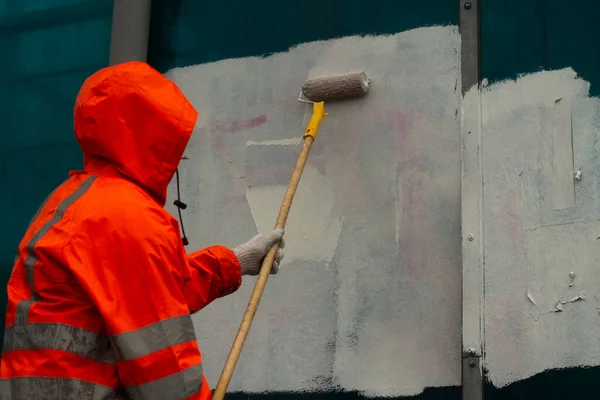 The painter paints with white paint. Paints graffiti with paint. Painting the wall. A worker paints over a vandalist inscription.
