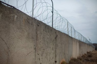 Dikenli tellerle. Bölgeyi çitle çevreleyen. Uzakta bir çit var. Tepesinde tel olan beton duvar..