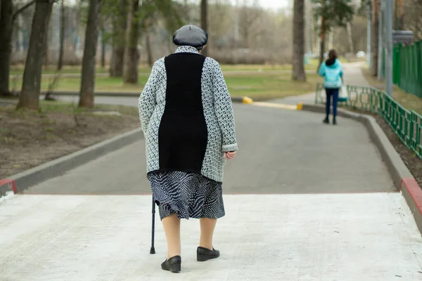 Elderly woman with a stick. Retired in Russia. Granny walks along the road. Shooting from the back.
