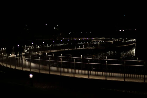 The path in the park at night. Illumination of the bridge near the water. Dark time in the city.
