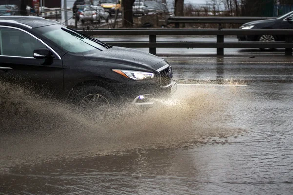 The car is driving through a puddle. Splashes from a puddle on the road. The car, dirty from precipitation, is driving along the highway. A large puddle on the road under the wheels of a car.