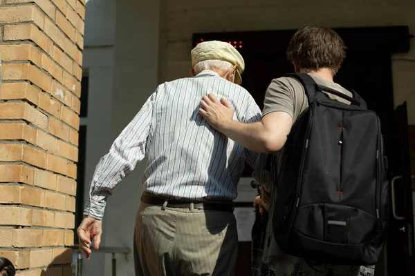 The grandson helps grandpa walk to the entrance to the hospital. A volunteer leads a pensioner to be vaccinated. A social worker accompanies an old man.