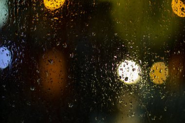 Cama yağmur damlaları damlıyor. Işıklı bulanık arkaplan. Koyu arkaplandaki camın dokusu. Yuvarlak şekilli bulanık ışıklar.