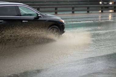 Araba bir su birikintisinin içinden geçiyor. Yoldaki su birikintisinden damlıyor. Sağanak yağıştan dolayı kirlenen araba otoyol boyunca sürüyor. Bir arabanın tekerleklerinin altında büyük bir su birikintisi.