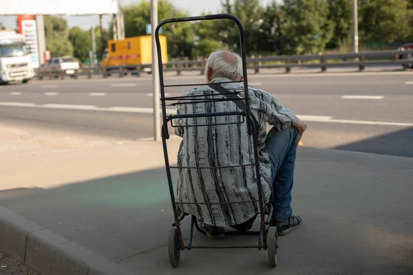 An old man at a bus stop. Grandpa is sitting on a cart. The pensioner is waiting for transport. The gray-haired man is tired. A man in the shade from the heat.
