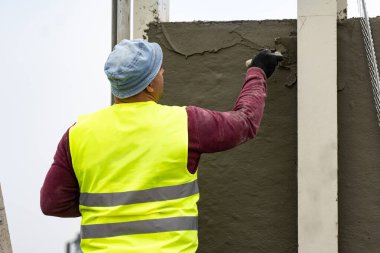 Yansıtıcı ceketli bir işçi inşaat alanının duvarına alçı yapıştırıyor.