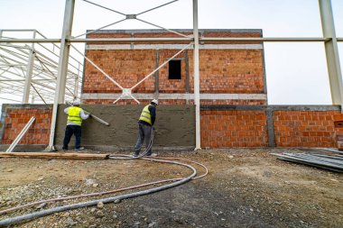 Yansıtıcı ceketli işçiler duvara alçı uyguluyorlar. Alçı duvarı yıkıyorum.