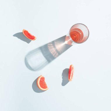 Bir bardak greyfurt suyu ve açık mavi arka planda taze greyfurt dilimleri. Asgari güneş ışığı estetiği.