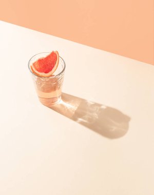 Parlak bej ve turuncu arka planda bir dilim taze greyfurtla bir bardak içki. Yaratıcı asgari yaz konsepti. Güneşli gün gölgesi.