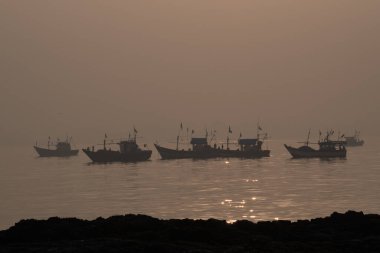 Gün doğarken deniz suyunda yüzen siluet botlar ön planda kayalarla birlikte deniz kıyısında