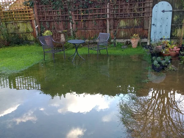 Wymondham Norfolk 'taki aile evinde şiddetli yağmur fırtınasından sonra sel basmış bahçenin manzarası gökyüzü ve bulut çimlerin üzerindeki su havuzuna yansıyor ve eski kapıya giden yolda ahşap arka planda duran metal koltuklar.