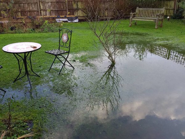 Landscape of flooded garden after heavy rain storms in family home in Wymondham Norfolk East Anglia uk with sky and cloud reflected in the pool of water on the grass lawn and path to old door with metal chairs table by wood fence background in Winter