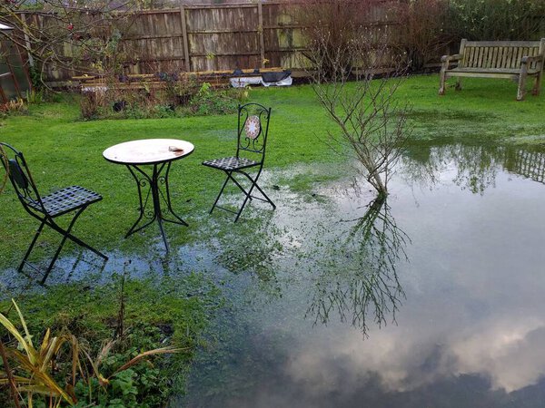 Landscape of flooded garden after heavy rain storms in family home in Wymondham Norfolk East Anglia uk with sky and cloud reflected in the pool of water on the grass lawn and path to old door with metal chairs table by wood fence background in Winter