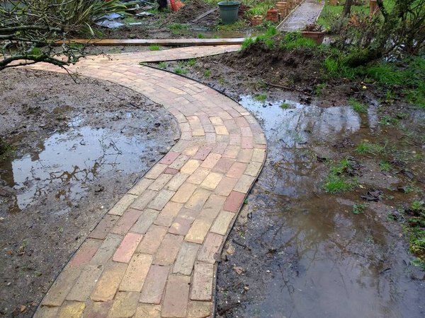 Close up of flooded garden path newly constructed from reclaimed ancient house bricks and laid on hardcore to create curving lane with the flood water forming pools wither side on the sodden earth and heavy clay soil ground