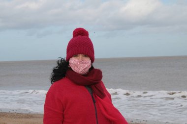 Kırmızı ceket atkısı ve şapkalı, siyah saçlı, Winterton Norfolk 'ta okyanus kıyısında güzel kumlu sahilde oturan Covid-19 virüslü deniz salgını nedeniyle kilitlenmiş bir kadın.