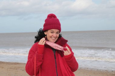 Kırmızı ceket atkısı ve şapkalı, siyah saçlı, maske takan, Winterton Norfolk 'un doğusundaki Kum Denizi kıyısında güzel bir sahile oturmuş bir kadın, Covid-19 virüs salgını nedeniyle İngiltere' de kilit altında tutuluyor.