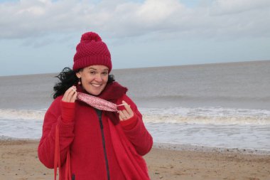 Kırmızı ceket atkısı ve şapkalı siyah saçlı bir maske takmak üzere olan bir kadın Winterton Norfolk 'un doğusundaki güzel kumlu sahil kıyısında Covid-19 virüs salgını denizaltı arka planında tutuluyor.