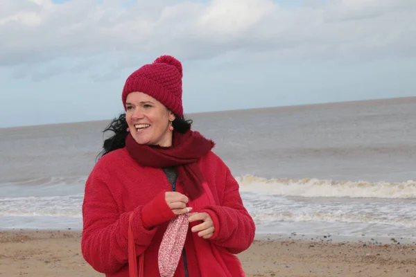 Close up of female about to put on a mask with black hair in red clothes of red jacket scarf and hat seated on seaside beautiful sandy beach by ocean sea shore in Winterton Norfolk east Anglia UK in lockdown for Covid-19 virus outbreak sea background