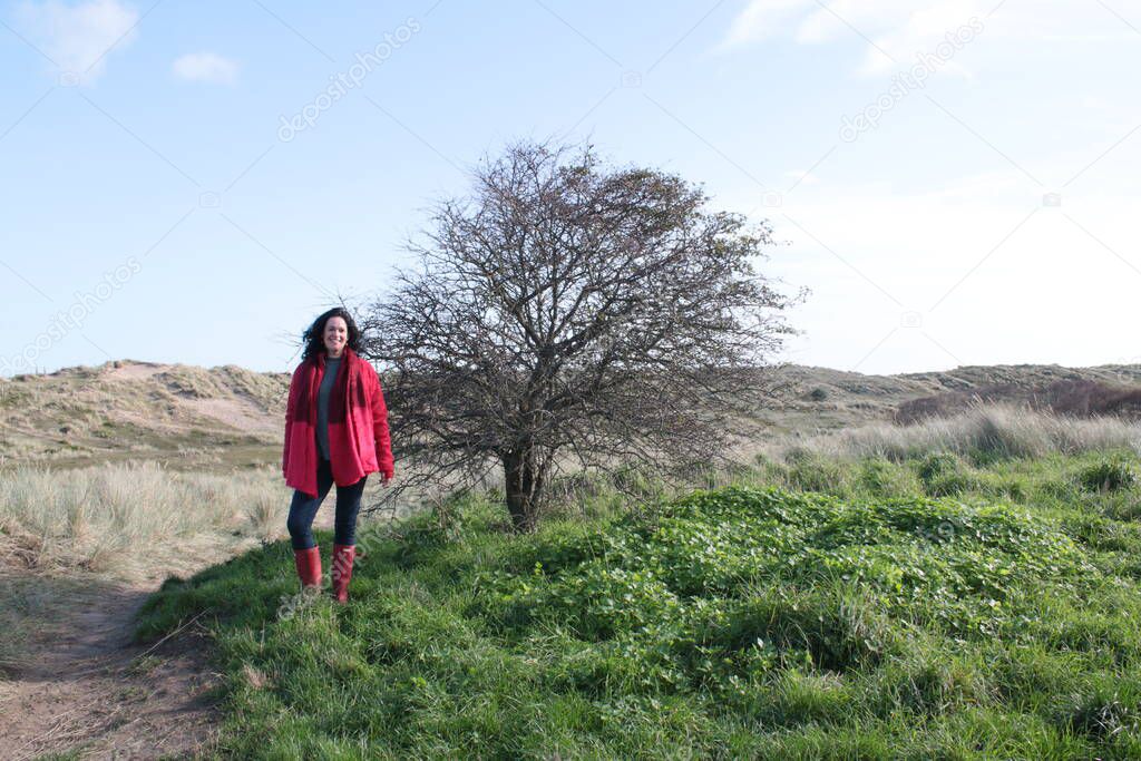 Attractive black haired female  middle aged in coast landscape standing smiling next to tree on grassy sand dunes by ocean fresh air wearing red scarf boots and coat with blue skies getting exercise during the covid-19 lockdown restrictions 2021